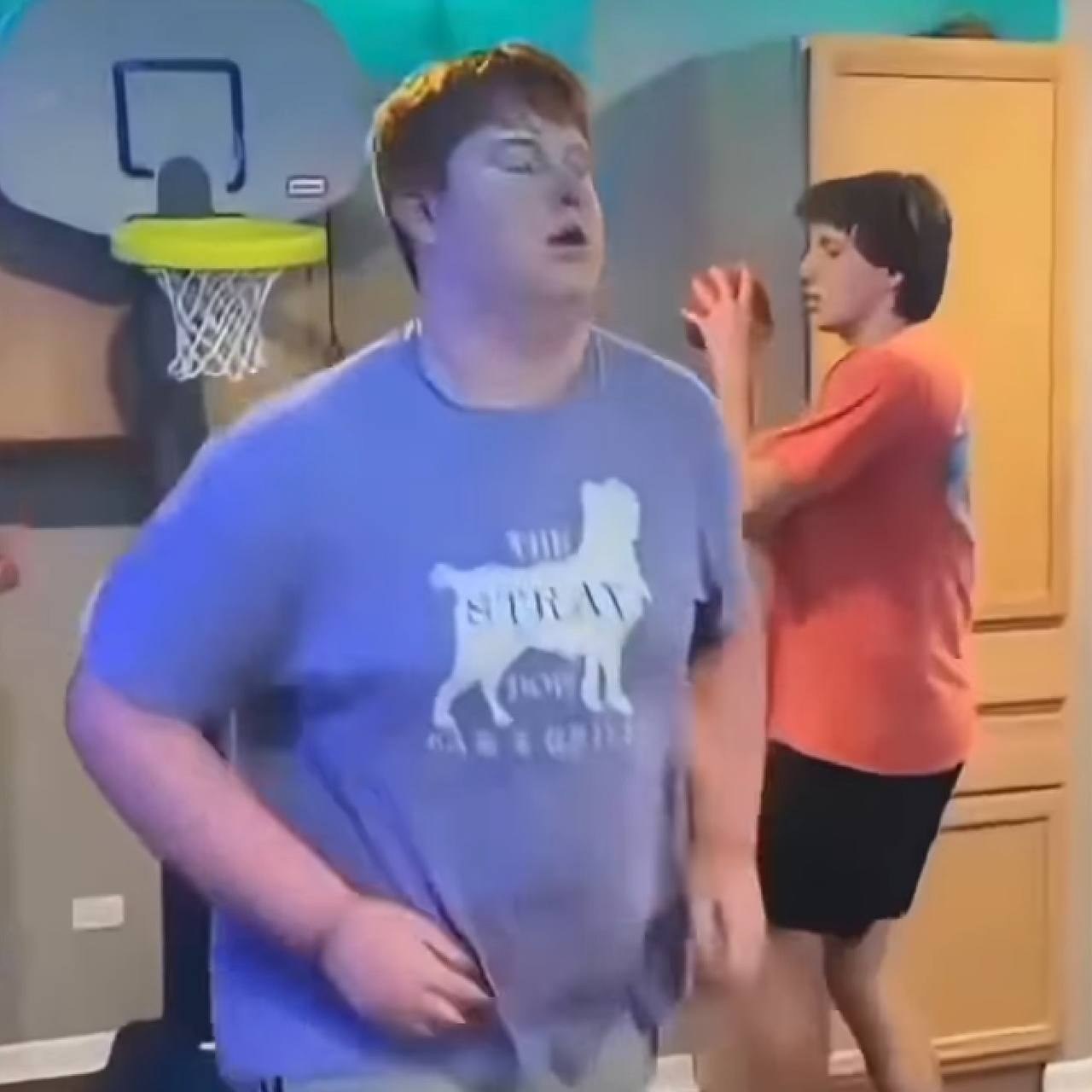 Grainy home-video frame of a red-haired guy walking away from a small wall-mounted basketball hoop in a living room